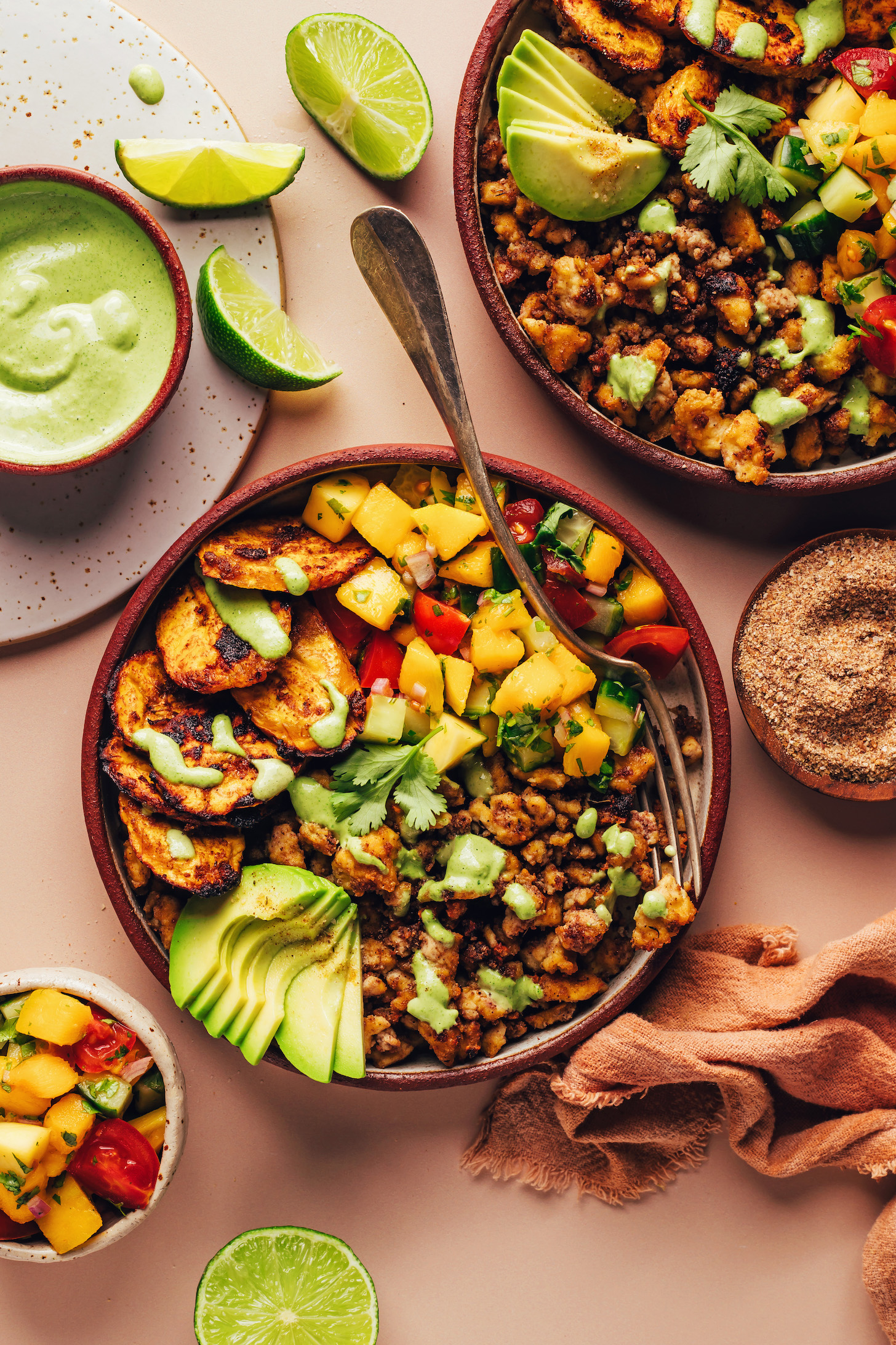 Small bowls of parsley tahini sauce and mango salad next to lime wedges and Jerk Tofu & Roasted Plantain Bowls