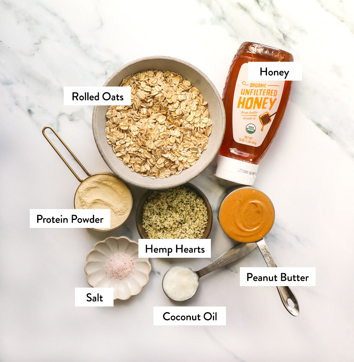 oats, protein powder, honey, peanut butter and hemp hearts labeled on a white surface.