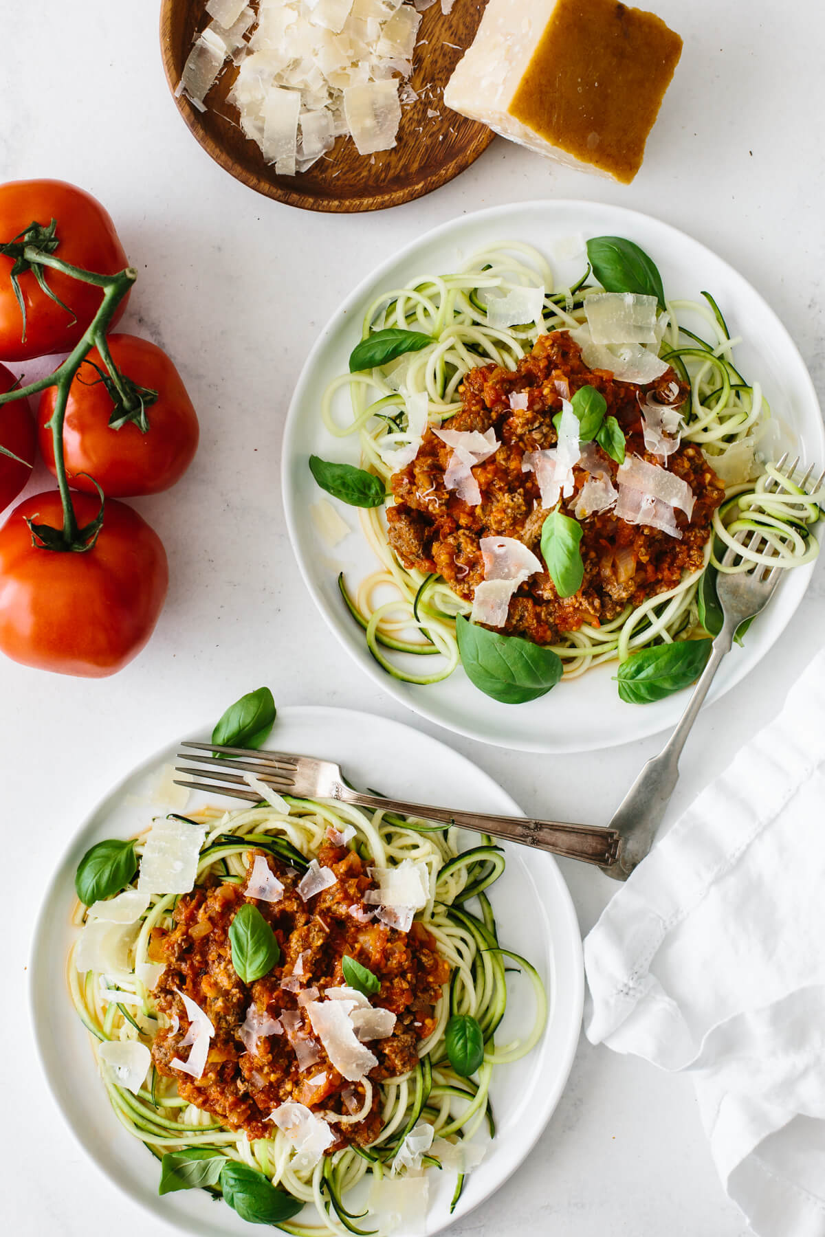 Two plates of zucchini noodle spaghetti bolognese, with tomatoes and parmesan on the side.