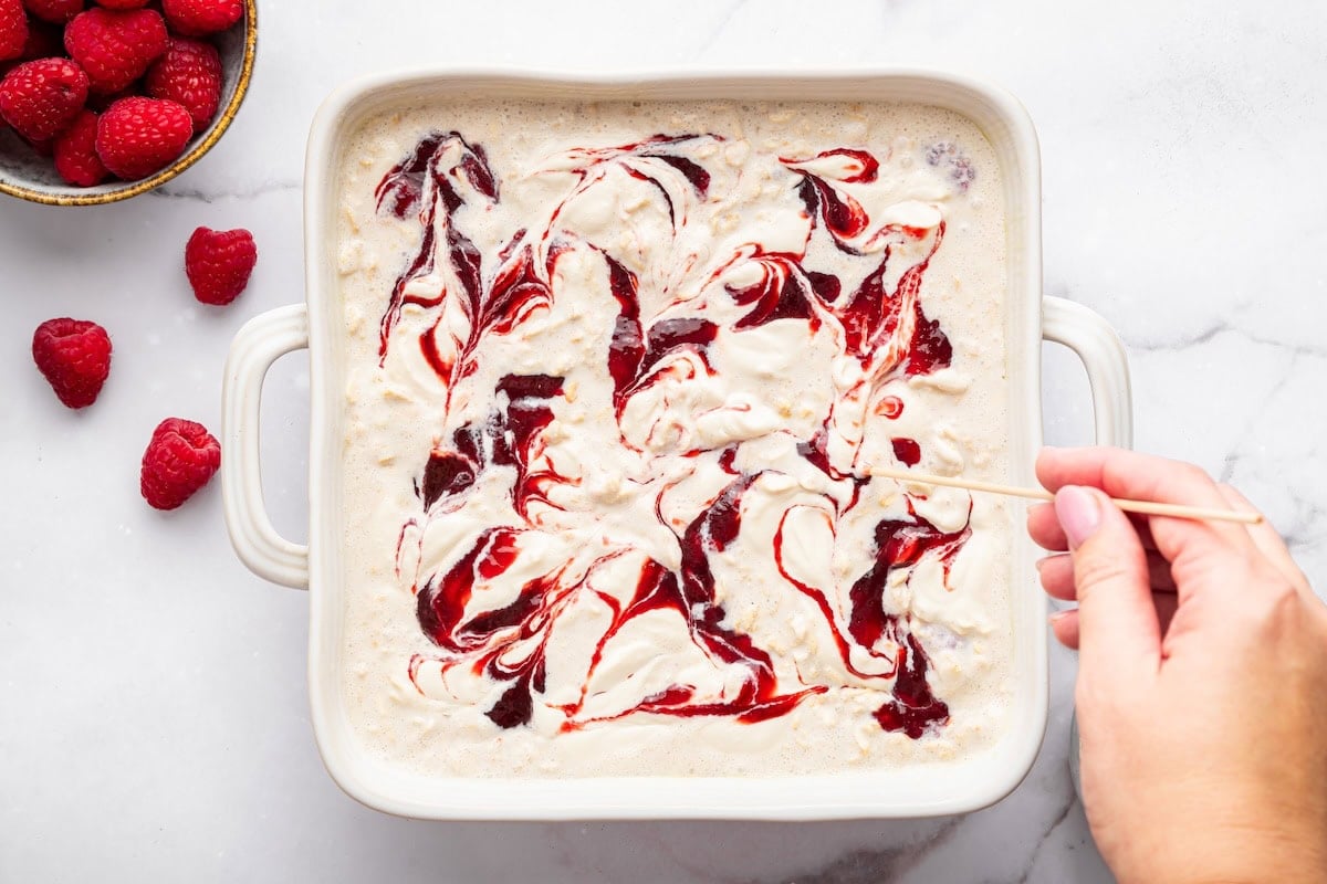 Unbaked oatmeal mixture in pan topped with raspberry jam and cottage cheese swirls.