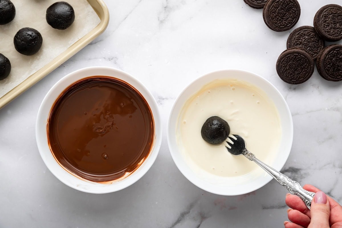 Oreo ball being dipped into melted white chocolate using a fork for coating.
