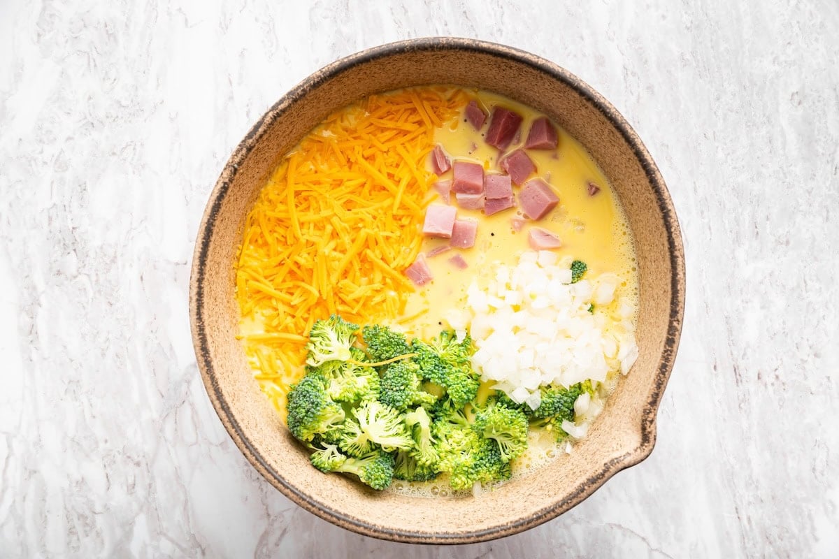 A large mixing bowl with eggs, shredded cheddar cheese, diced ham, broccoli, and onion.