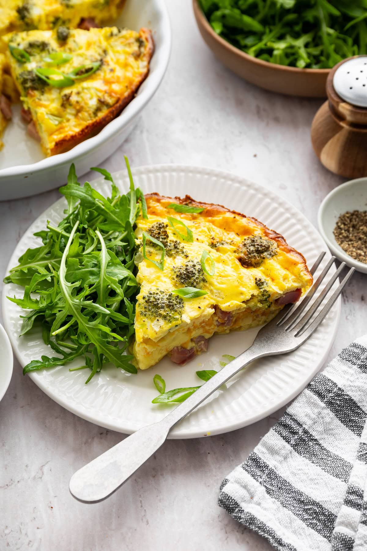 A slice of crustless quiche on a white plate with a fork and an arugula salad on the side.