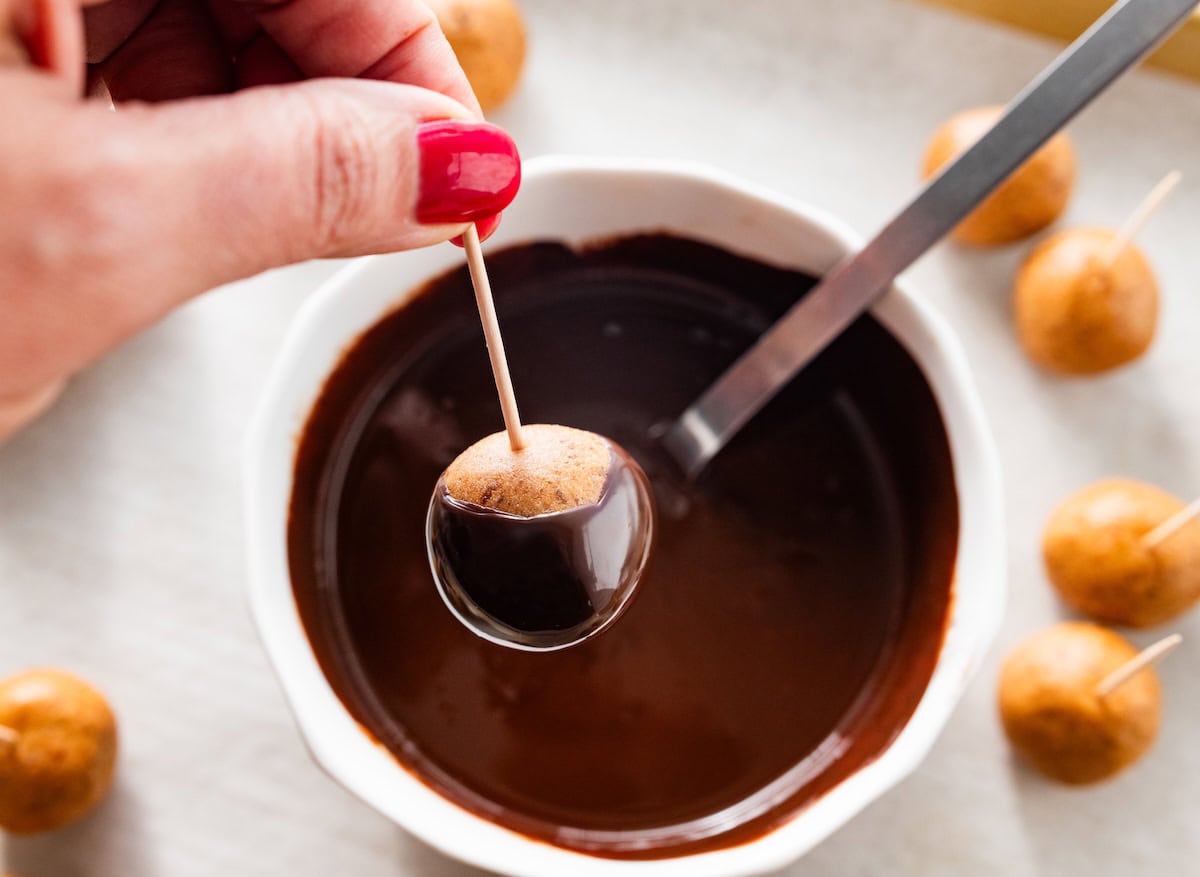 A peanut butter buckeye ball being dipped halfway into melted dark chocolate with a toothpick.