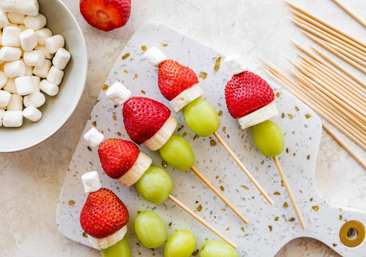 Grinch fruit kabobs scattered on a marble cutting board with bowls of fruit and toothpicks nearby.