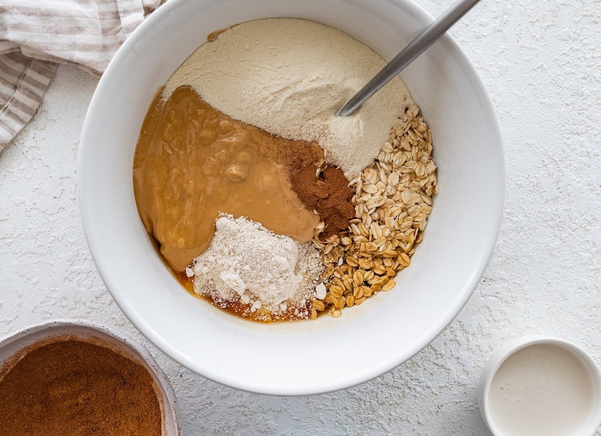 ingredients in Mixing bowl Mixing bowl filled with oats, oat flour, cashew butter, protein powder and cinnamon before stirring.