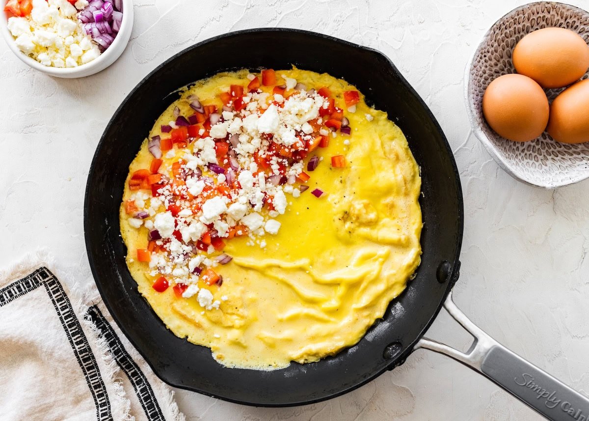 Omelette in a pan topped with chopped peppers, red onion and crumbled feta before folding.