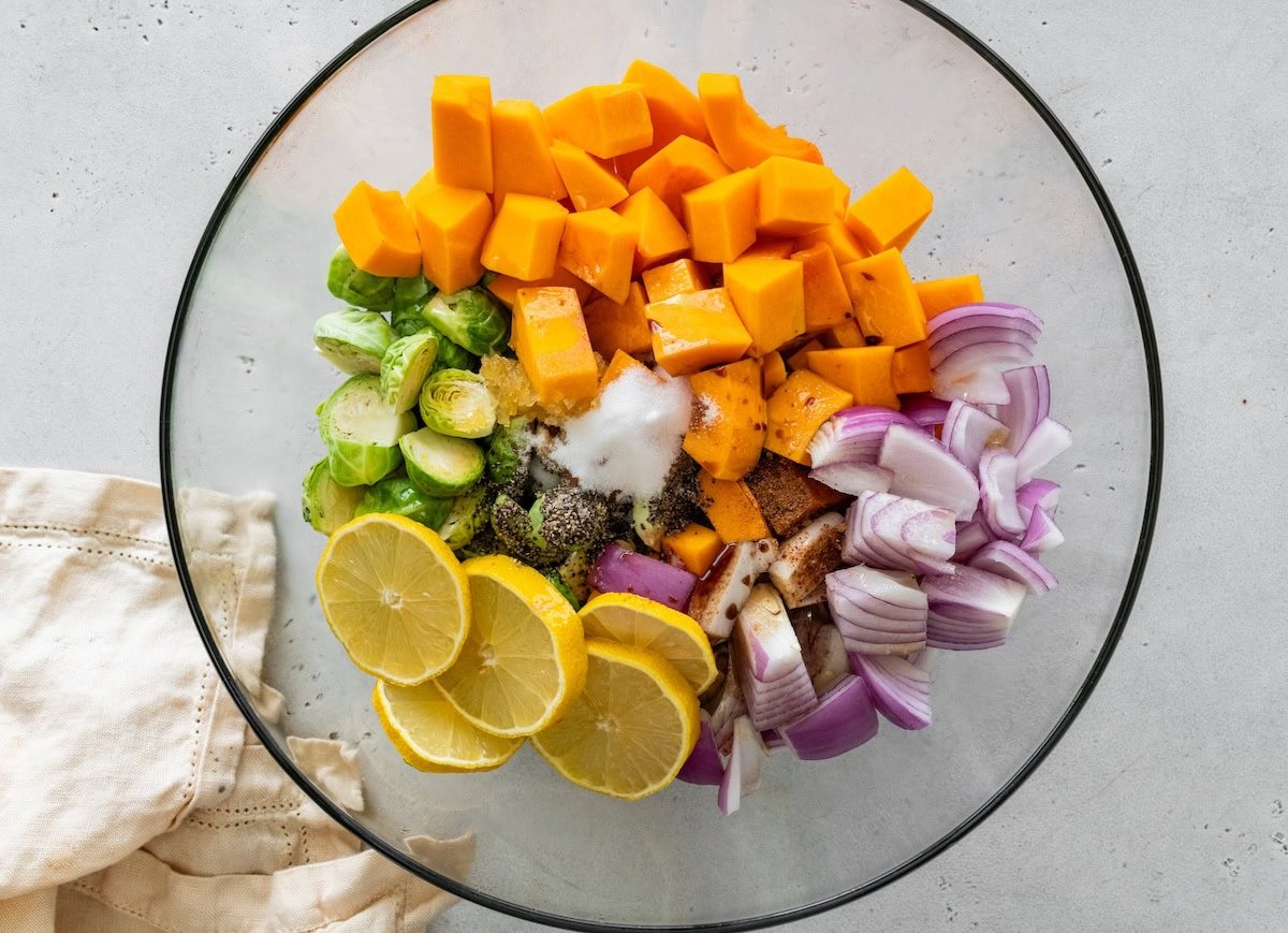 Chopped butternut squash, red onion, brussels sprouts and lemon in a glass mixing bowl sprinkled with seasonings and oil.