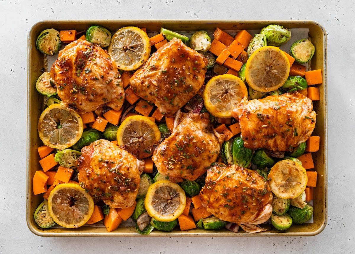 Sheet pan chicken thighs surrounded by roasted Brussels sprouts, squash, and sliced lemon.
