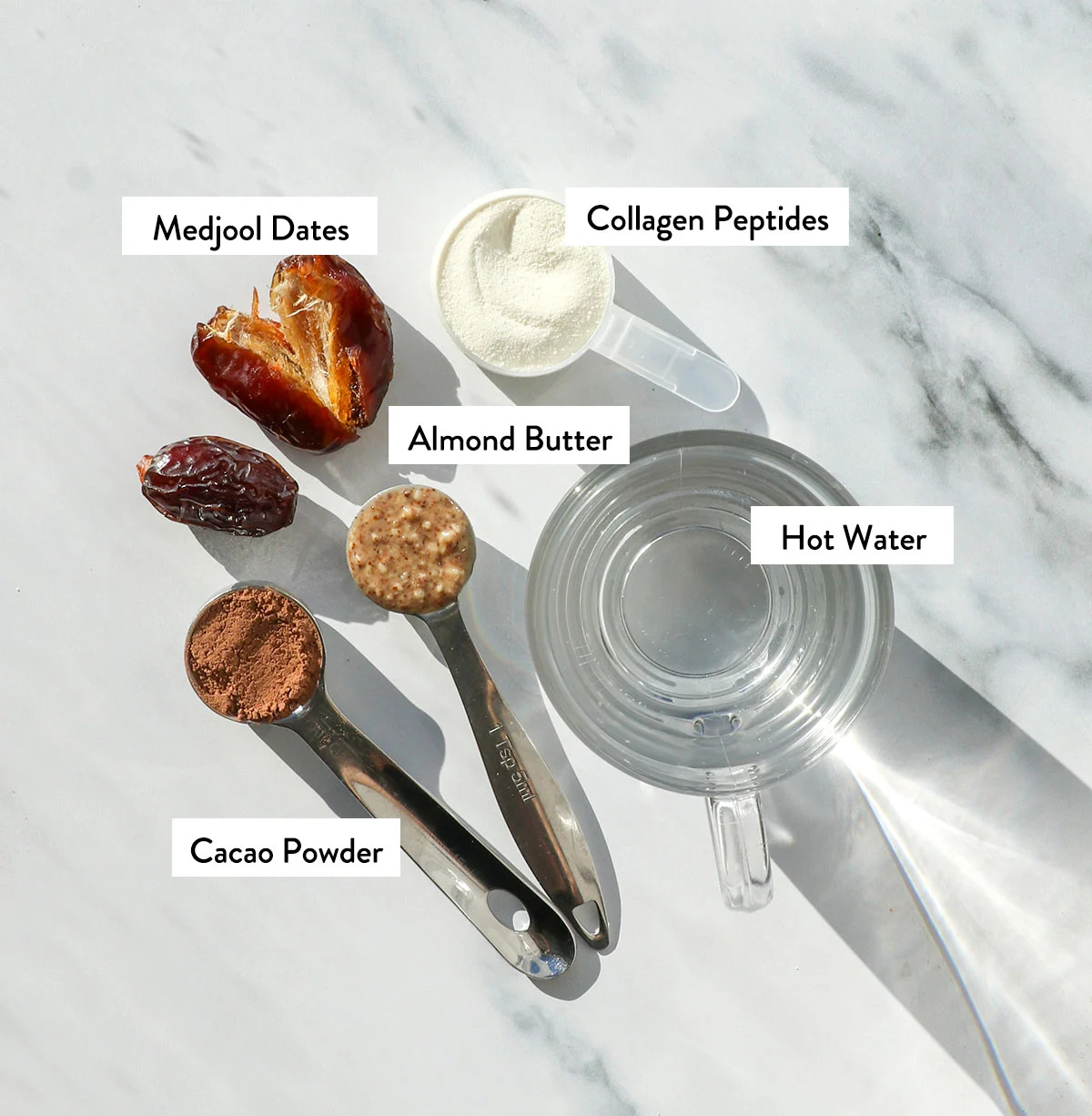 dates, almond butter, collagen, water, and cacao powder labeled on a white surface. 