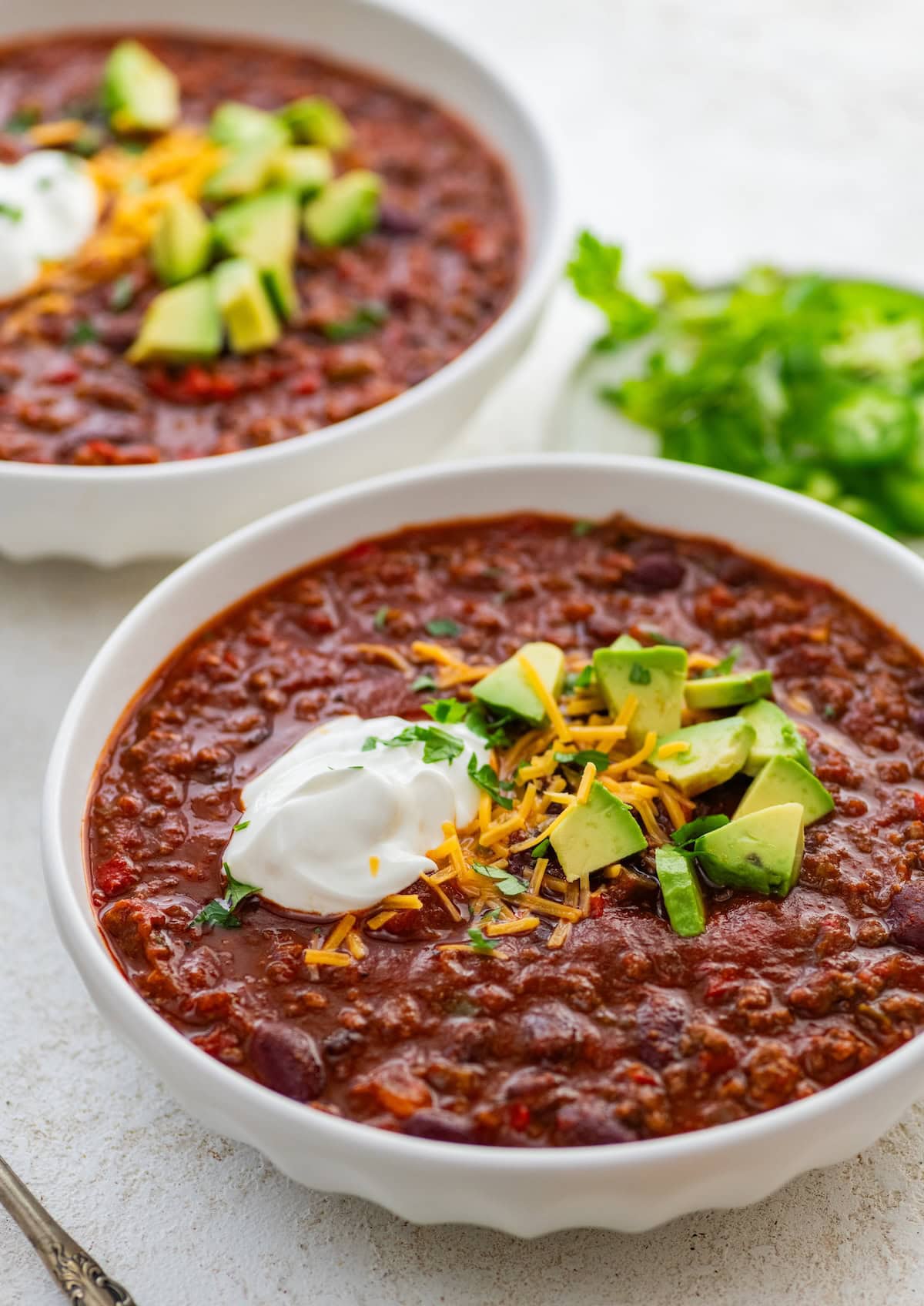 Two bowls of slow cooked beef and bean chili topped with avocado, cheese, and sour cream.