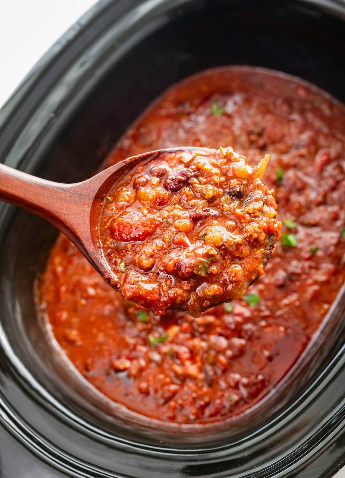 A wooden spoonful of slow cooked beef and bean chili held over a slow cooker.