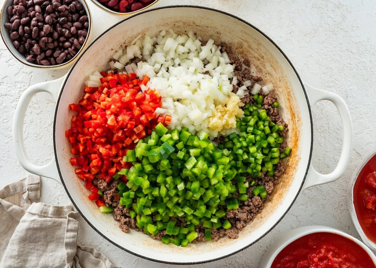 Diced red bell pepper, green bell pepper, onion, jalapeno, and garlic in a large pot with cooked ground beef.