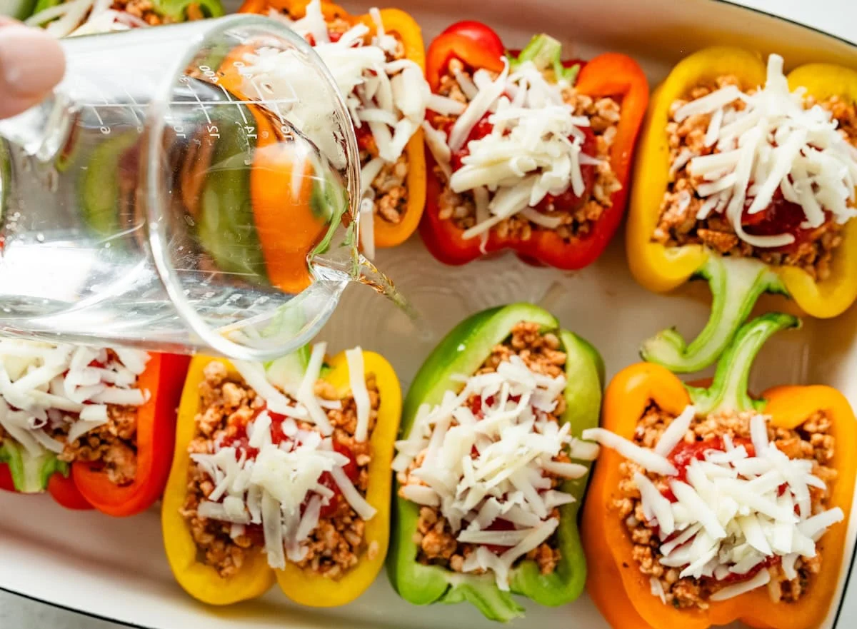 Water being poured into a pan of turkey stuffed peppers with shredded cheese on top.