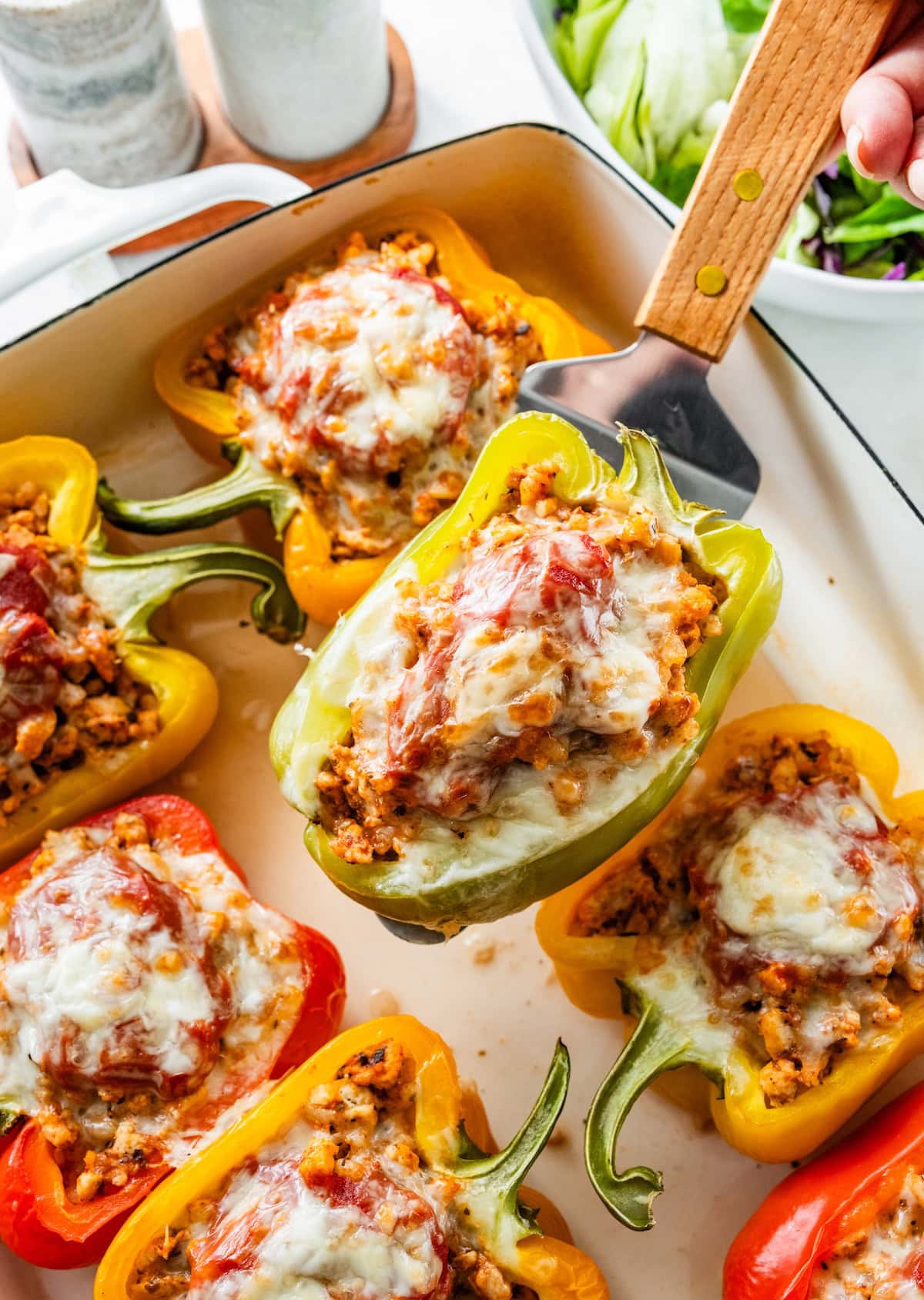 A turkey stuffed pepper on a spatula being held over a pan of more stuffed peppers.