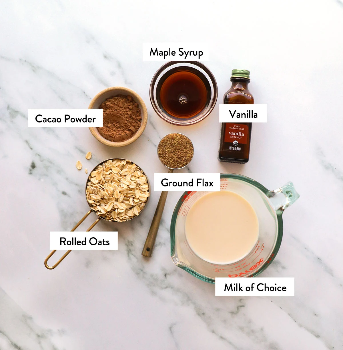 rolled oats, milk, cacao powder, maple syrup, and vanilla extract labeled on a white surface. 