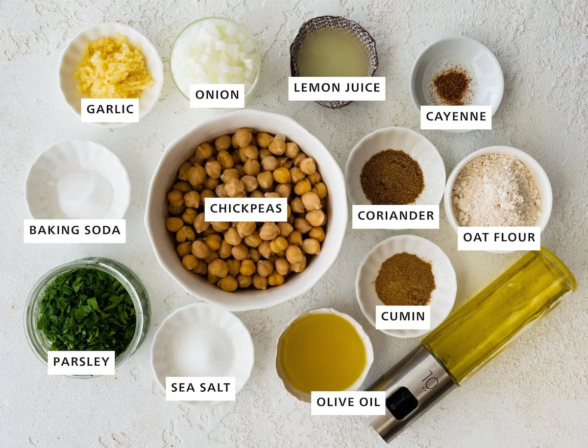 Ingredients portioned in small bowls for baked falafel including: chickpeas, cumin, coriander, oat flour, olive oil, sea salt, parsley, baking soda, garlic, onion, lemon juice, and cayenne.