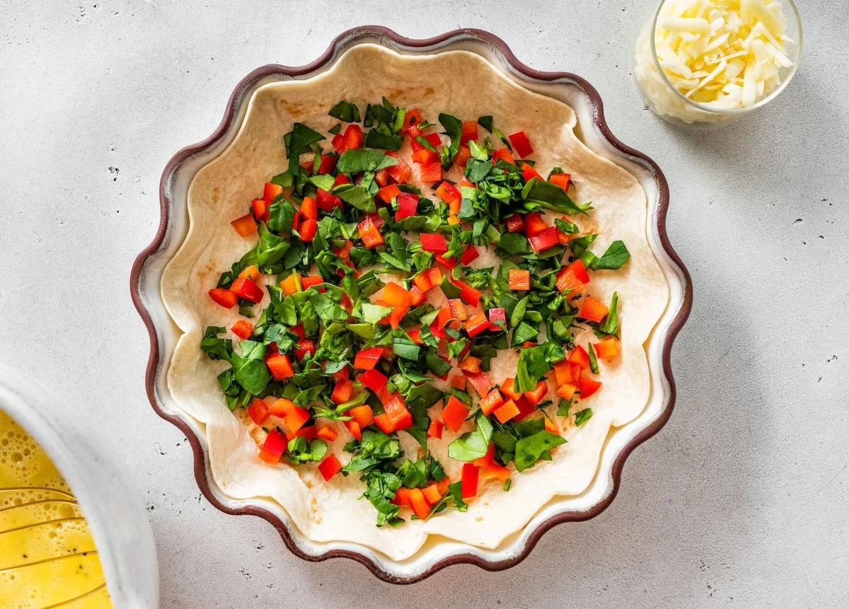 Flour tortilla pressed into pie dish topped with chopped spinach and red bell pepper.