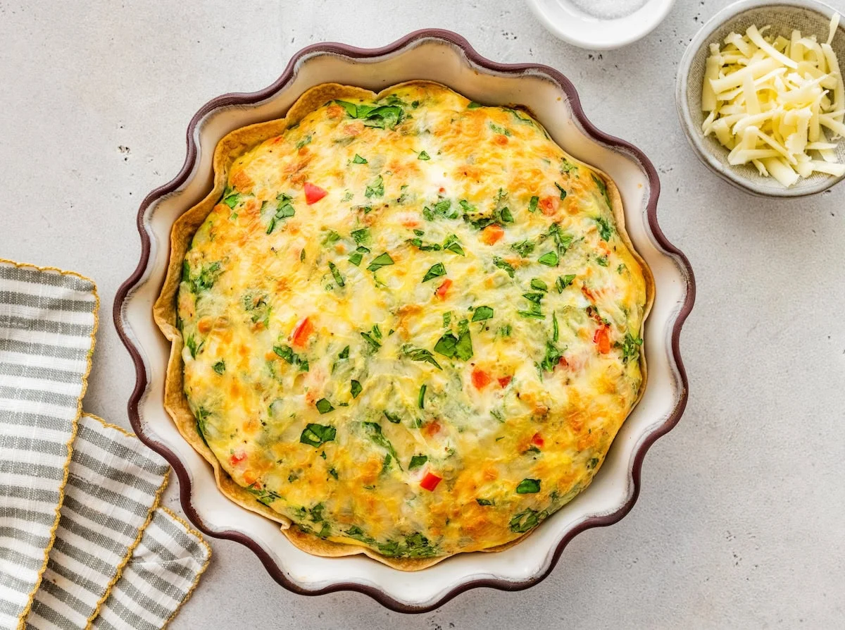 High-protein tortilla quiche baked with eggs, spinach, peppers, and cheese in fluted pie dish.
