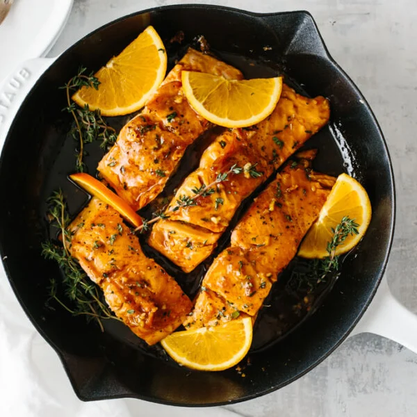 Four salmon filets in a pan with orange slices and thyme on top.