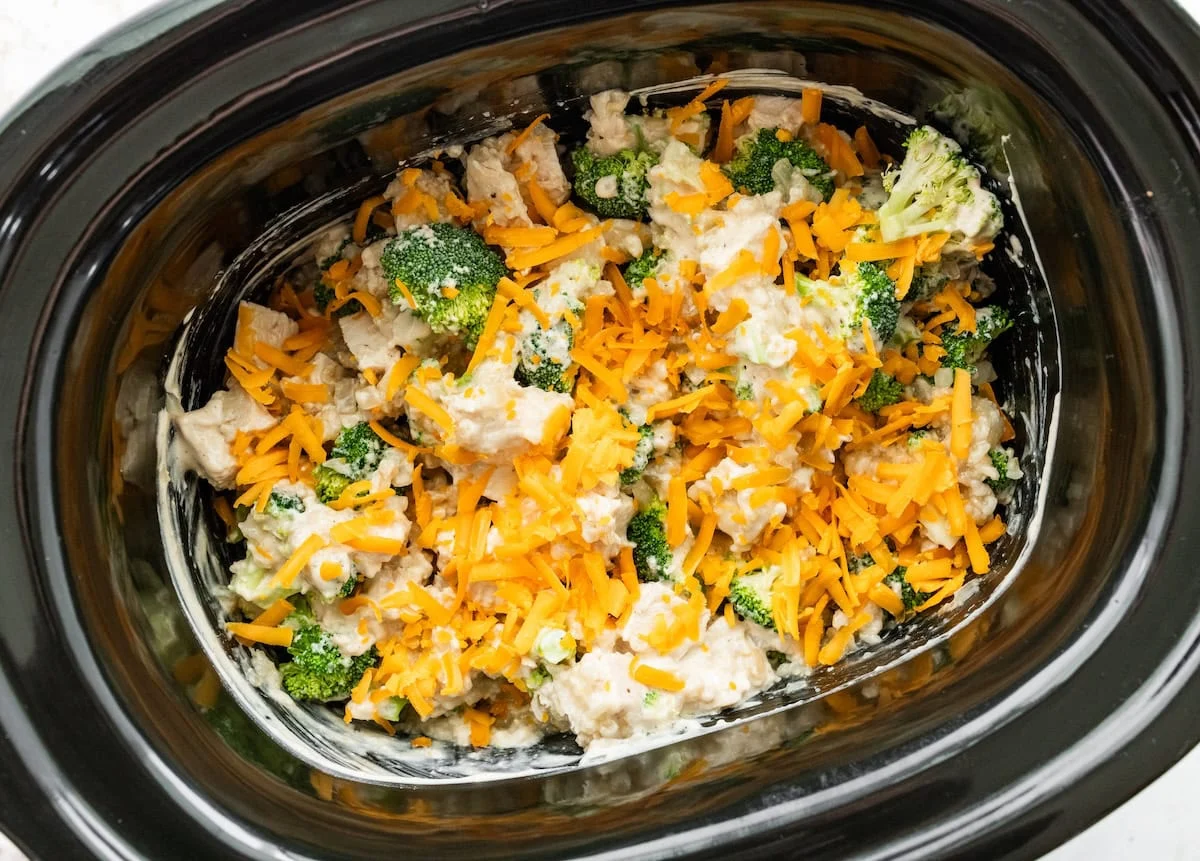 Shredded cheddar cheese added to a slow cooker over broccoli, chicken, rice, onion, garlic, chicken broth, and spices.