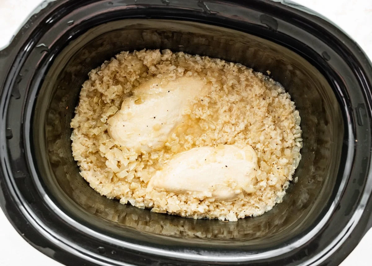 Cooked chicken breasts in a slow cooker with chicken broth, diced onion, minced garlic, brown rice, ground black pepper, and salt.
