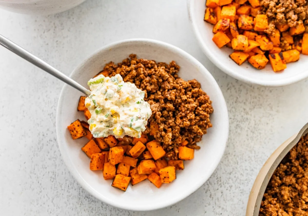 A white bowl with ground beef, diced sweet potato, and topped with a creamy street corn mixture.