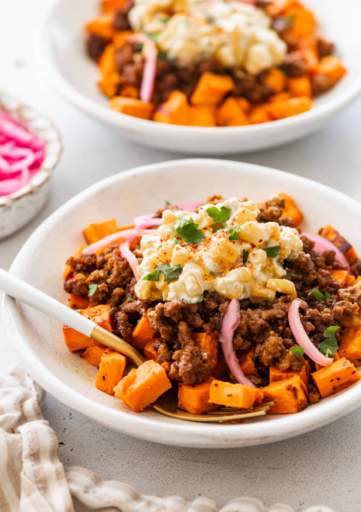 Roasted sweet potatoes in a white bowl topped with cooked ground beef, street corn salsa, pickled red onions, cilantro and a drizzle of hot honey.