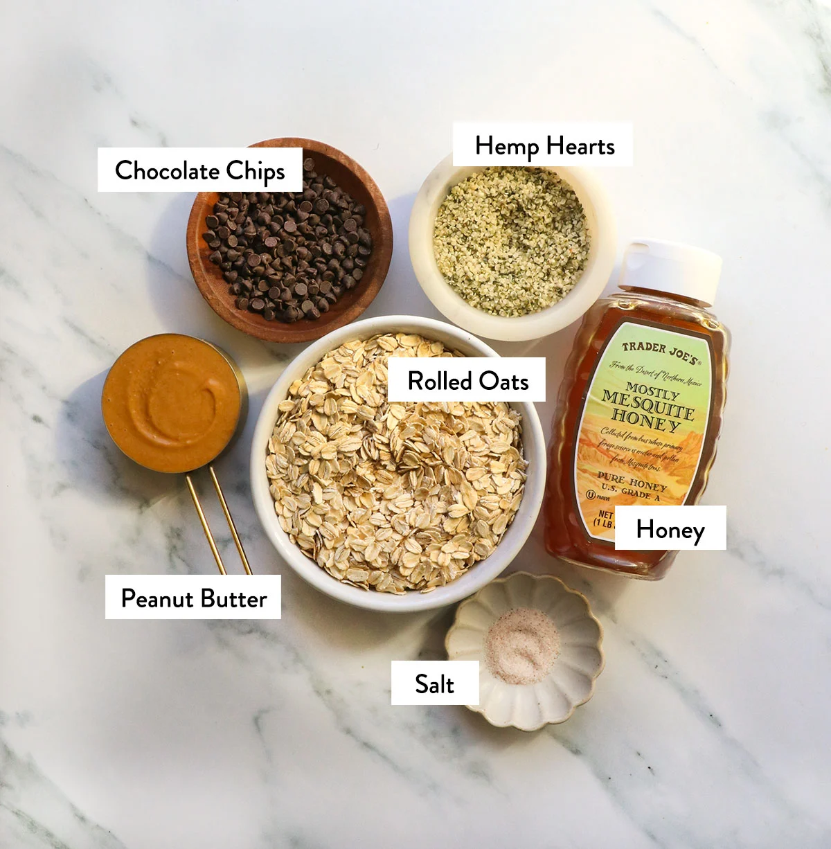 bowl of oats, honey, peanut butter, hemp hearts, and chocolate chips labeled in bowls.