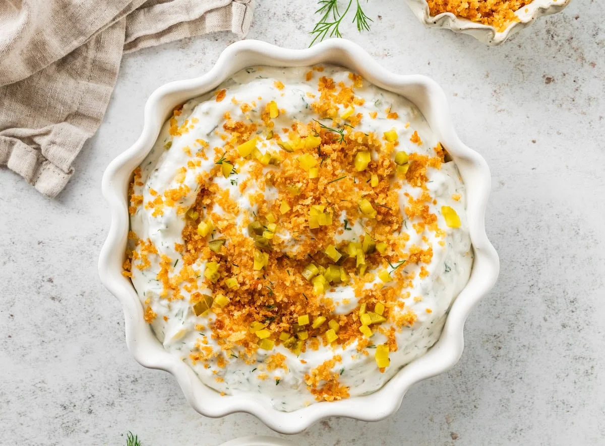 A white bowl with a high protein fried pickle dip. The dip has crispy breadcrumbs, diced pickle, and fresh dill on top.