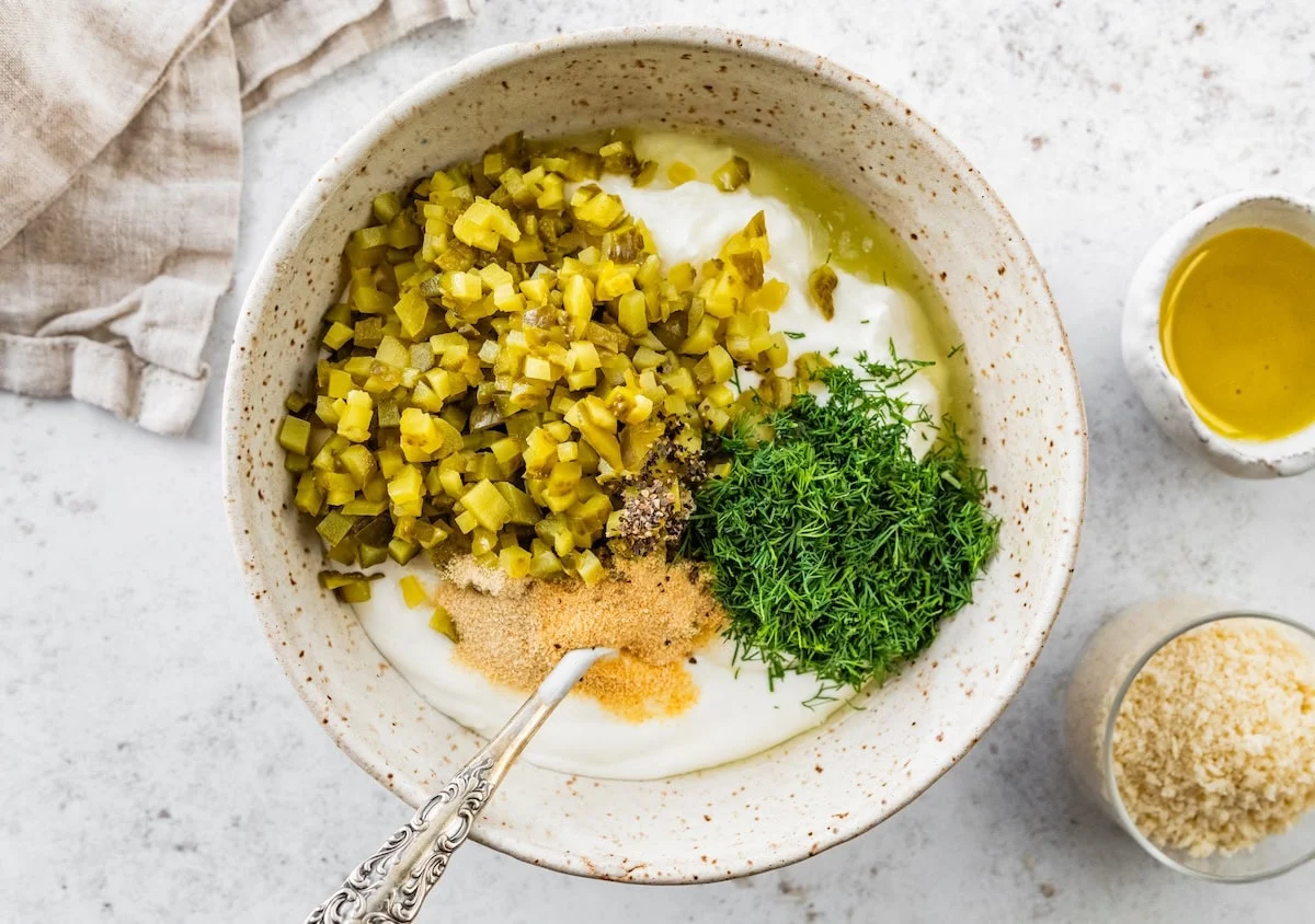 A large mixing bowl with greek yogurt, cream cheese, diced pickle, fresh dill, garlic powder, and onion powder.