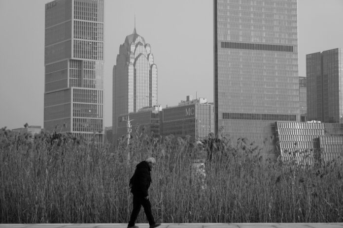 A person walks past tall buildings and reeds.
